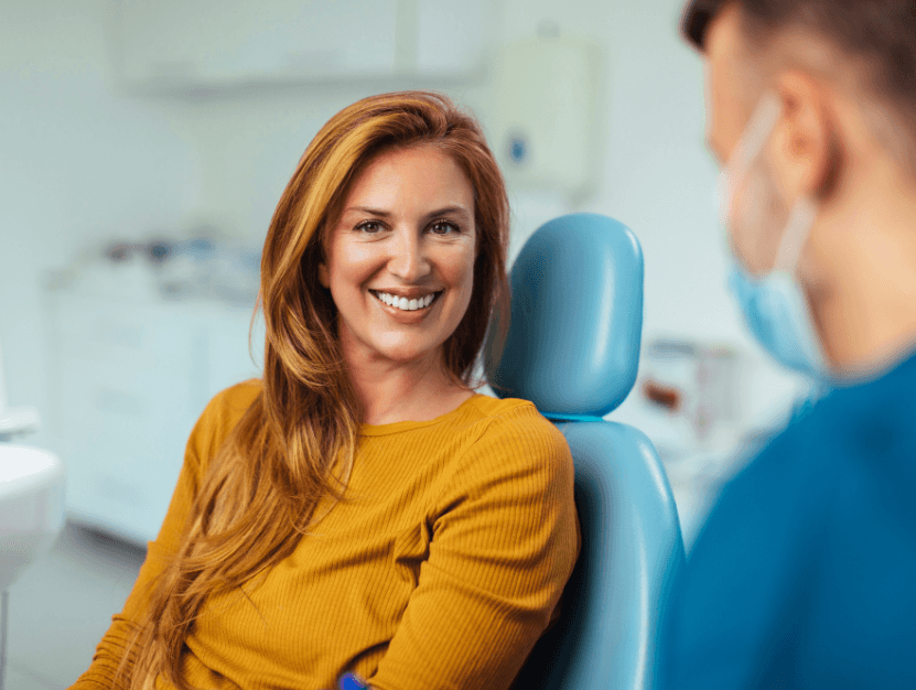 woman with ginger / auburn hair sitting on a dentists chair smiling at the camera with her teeth showing